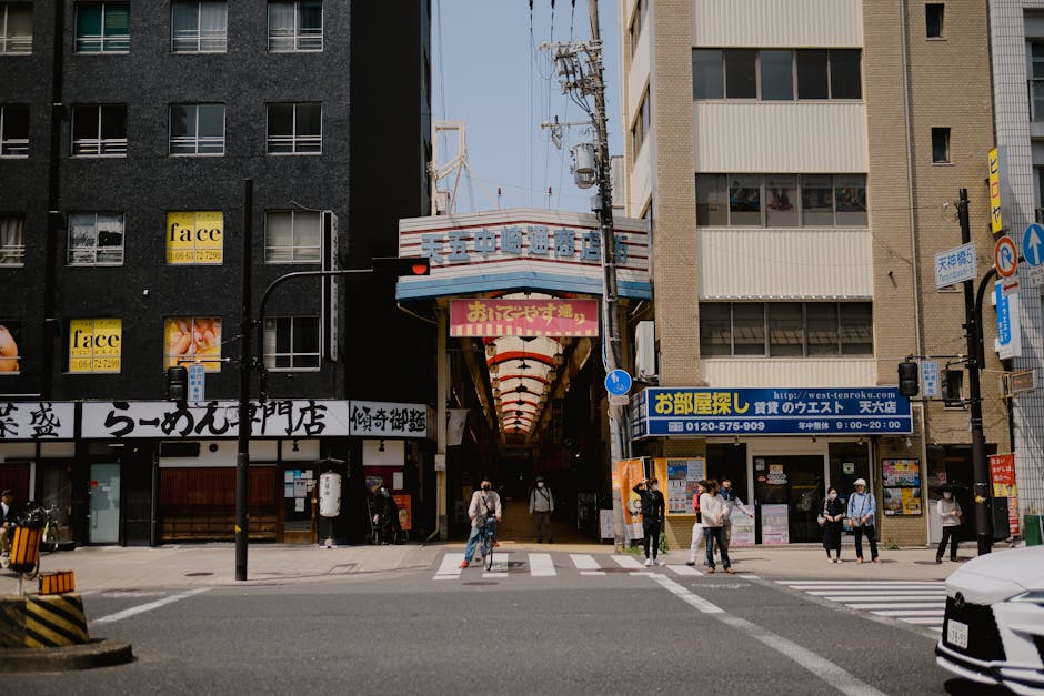 Street view of bustling Osaka with people crossing, shops, and arcade entrance.