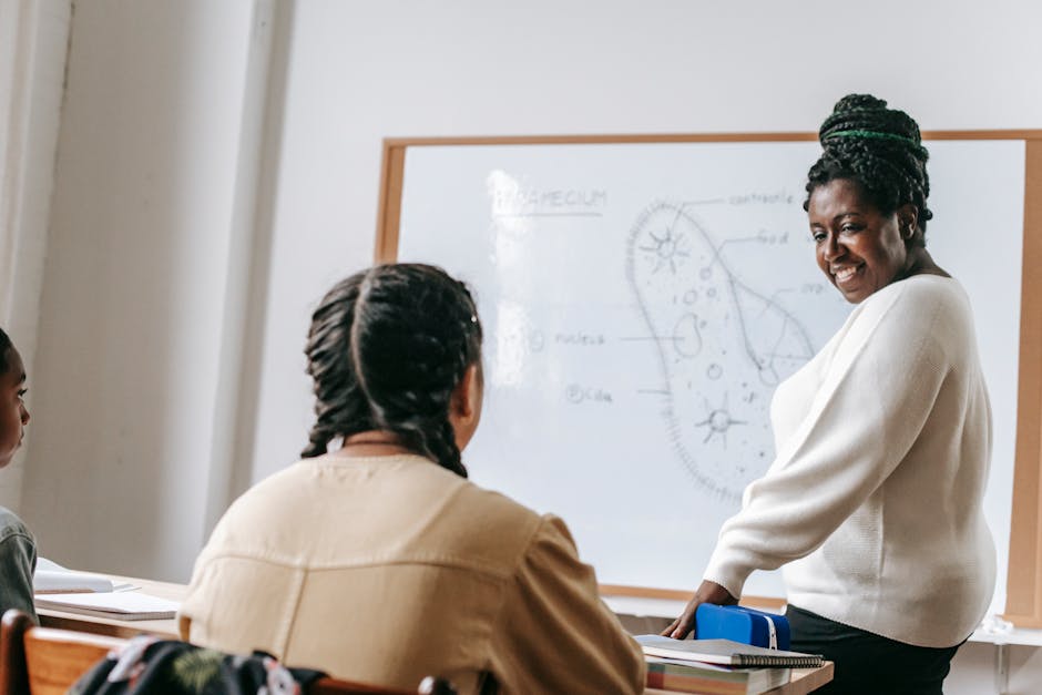 Side view of African American female teacher explaining material while standing near whiteboard and looking at teenage girl during biology lesson