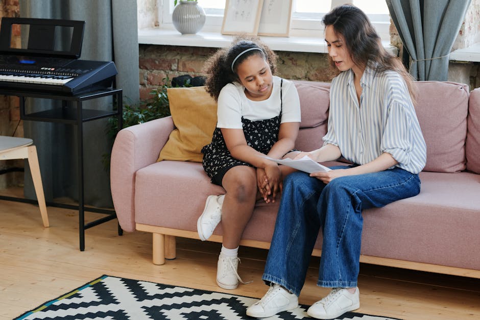 A woman tutors a girl in music theory on a sofa next to a synthesizer.