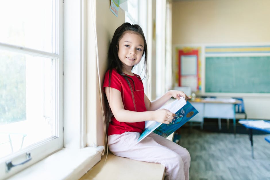 A young girl smiles while reading a book by the window in a bright classroom.