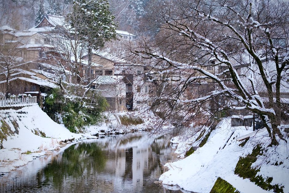 A picturesque winter view of snow-covered trees and traditional houses by a river in Takayama, Japan.