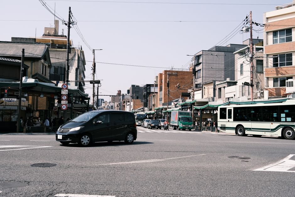 Street view of a bustling intersection in Kyoto, Japan. Features cars and buses in a lively urban setting.