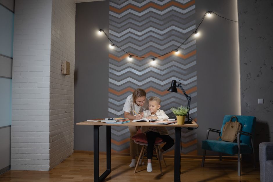 A mother assists her young son with homework at a desk in a cozy indoor setting.