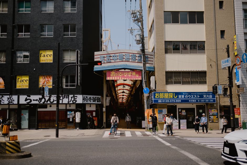 Street view of bustling Osaka with people crossing, shops, and arcade entrance.