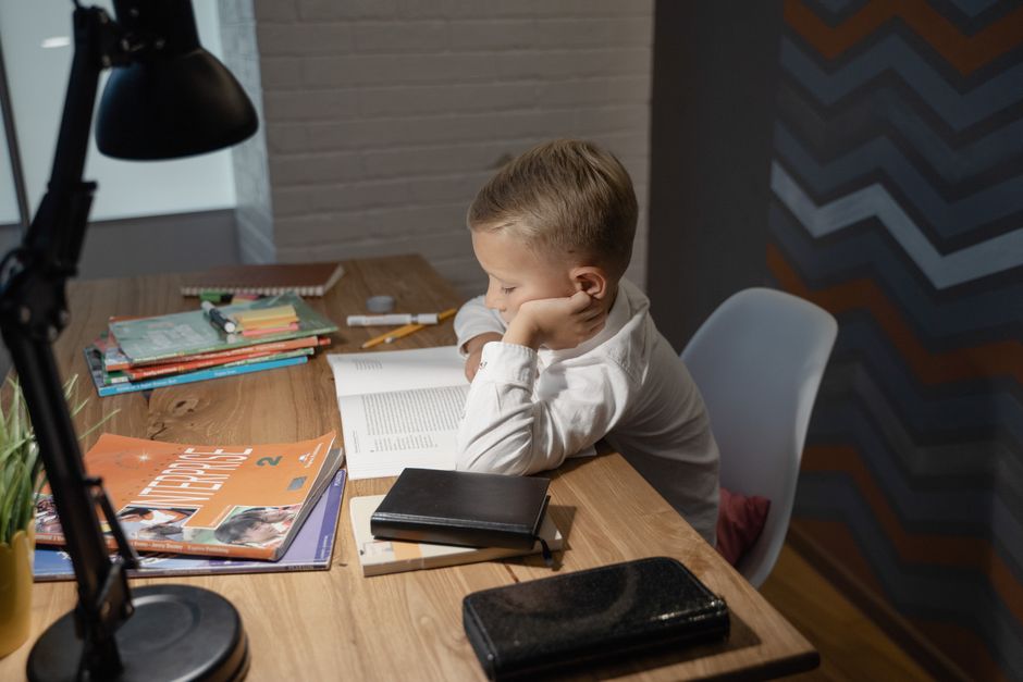 A young boy is reading a book at a desk with study materials and a desk lamp indoors.